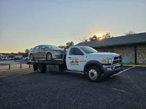 White Dodge Ram flatbed tow truck hauling a silver Lincoln sedan in Fort Worth, TX, with "Quick & Slick Towing" business decal and 469-427-7900 phone number, parked in front of a stone-faced building during a golden sunrise.